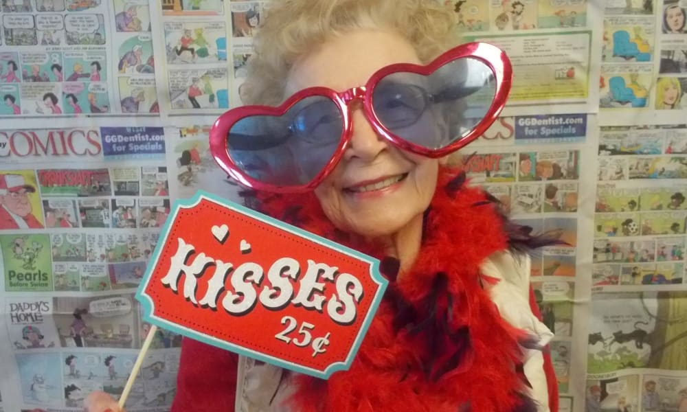 An elderly woman with a joyful expression wears oversized, heart-shaped sunglasses and a red feather boa. She holds a whimsical sign reading 
