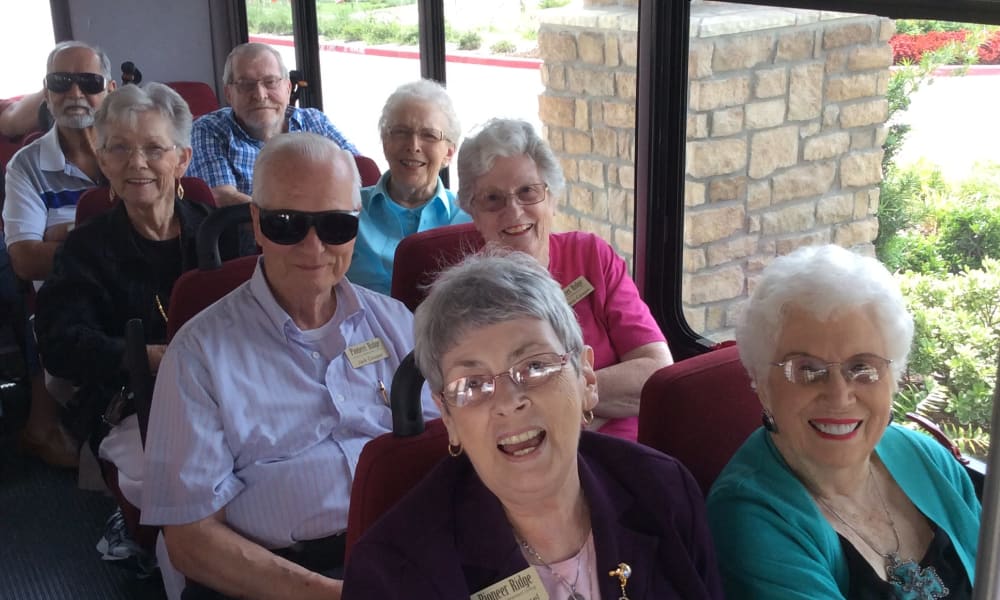 A group of smiling elderly individuals sits together in a bus, ready for a trip. Some wear sunglasses and name tags. They appear cheerful and are dressed in casual attire. The bus interior is filled with red seats, and a glimpse of the outside greenery is visible through the windows.