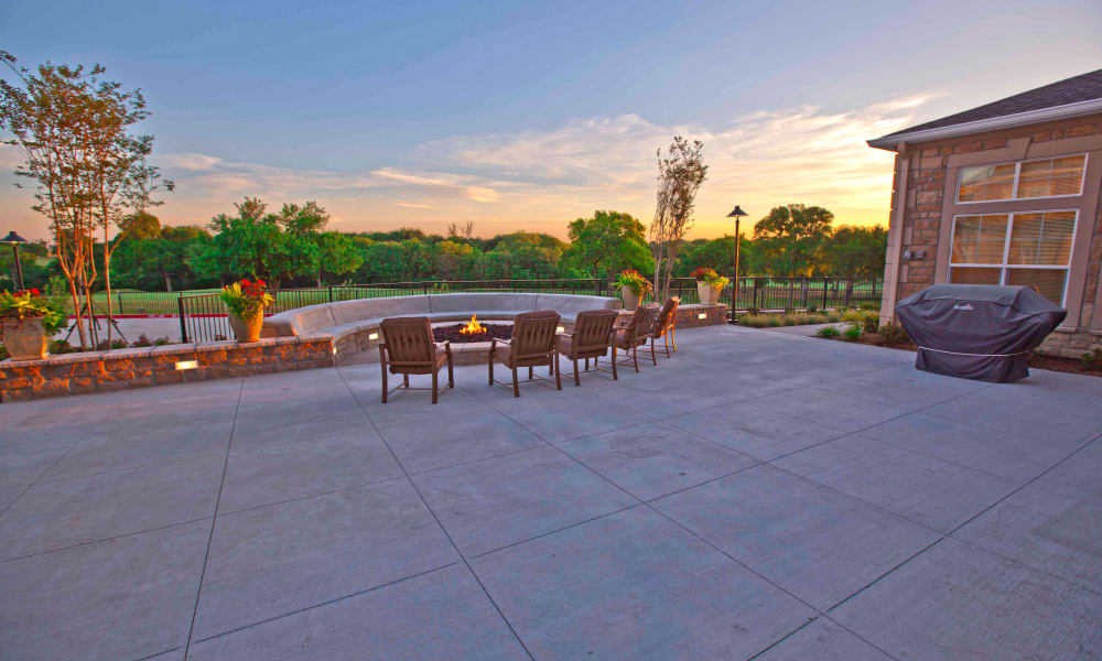 A spacious backyard with a paved patio featuring several wooden chairs around a fire pit. There is a grill covered with a black cover and lush green trees in the background. The sun is setting with a vibrant sky, casting a golden light over the scene.