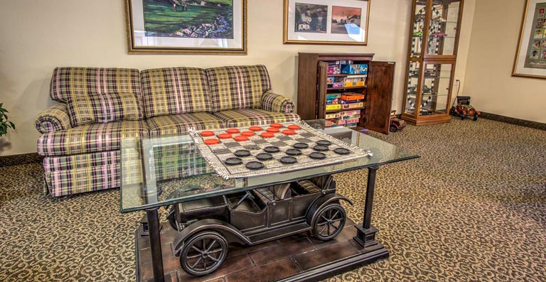 A cozy room with a plaid sofa, two framed pictures on the wall, a glass coffee table with a checkerboard game, and a wooden shelf displaying board games. The table has a metal model of a vintage car as its base. The floor is carpeted.