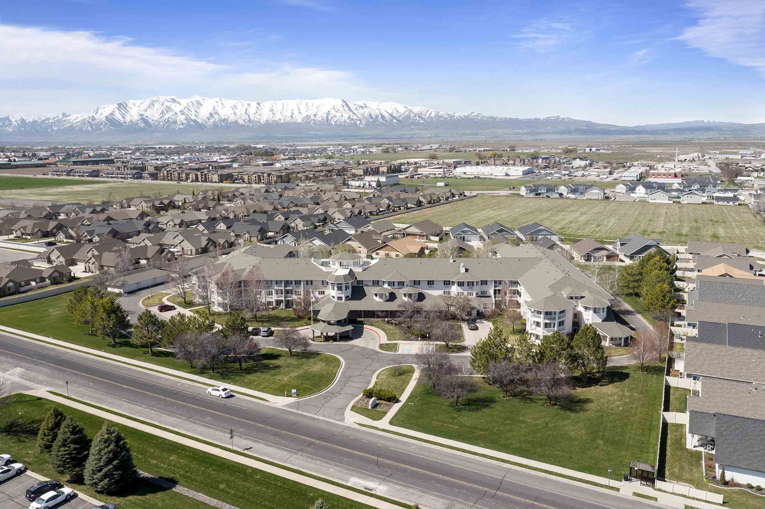 Aerial view of a suburban neighborhood with rows of houses and apartment buildings surrounded by green spaces. Snow-capped mountains are visible in the background under a clear blue sky. A main road runs through the foreground of the image.