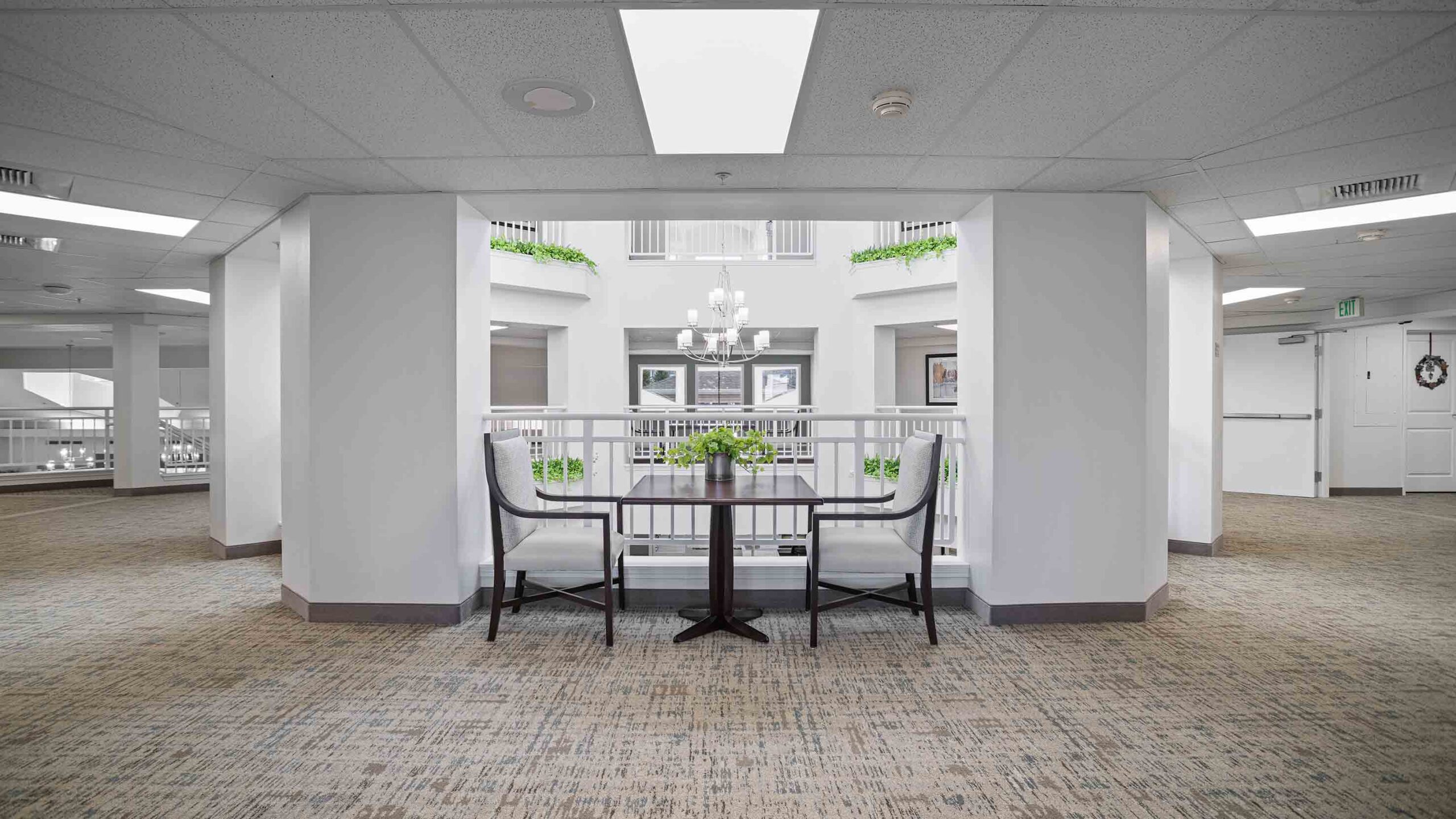 A modern indoor seating area featuring a small table with a plant and two upholstered chairs under bright overhead lighting. The area is surrounded by white walls and carpeted floors, with a railing and decorative plants in the background.