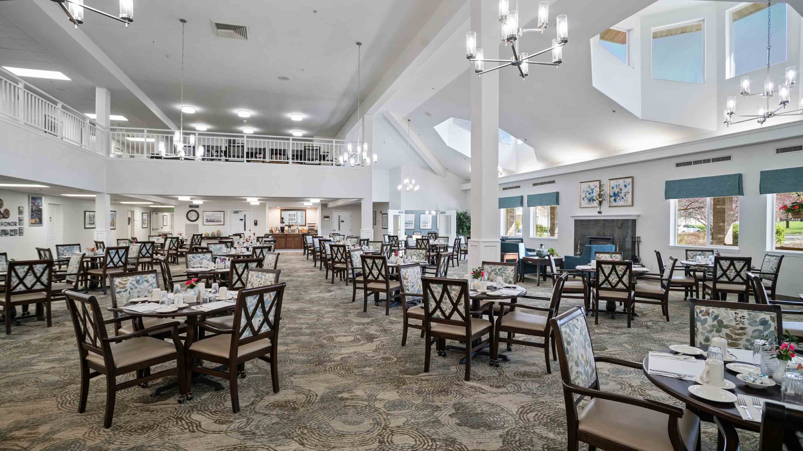 A spacious dining room with high ceilings, chandeliers, and tall windows. It features numerous tables set with cutlery, plates, and floral centerpieces, surrounded by cushioned chairs. A second-floor balcony overlooks the area, enhancing the open and airy ambiance.