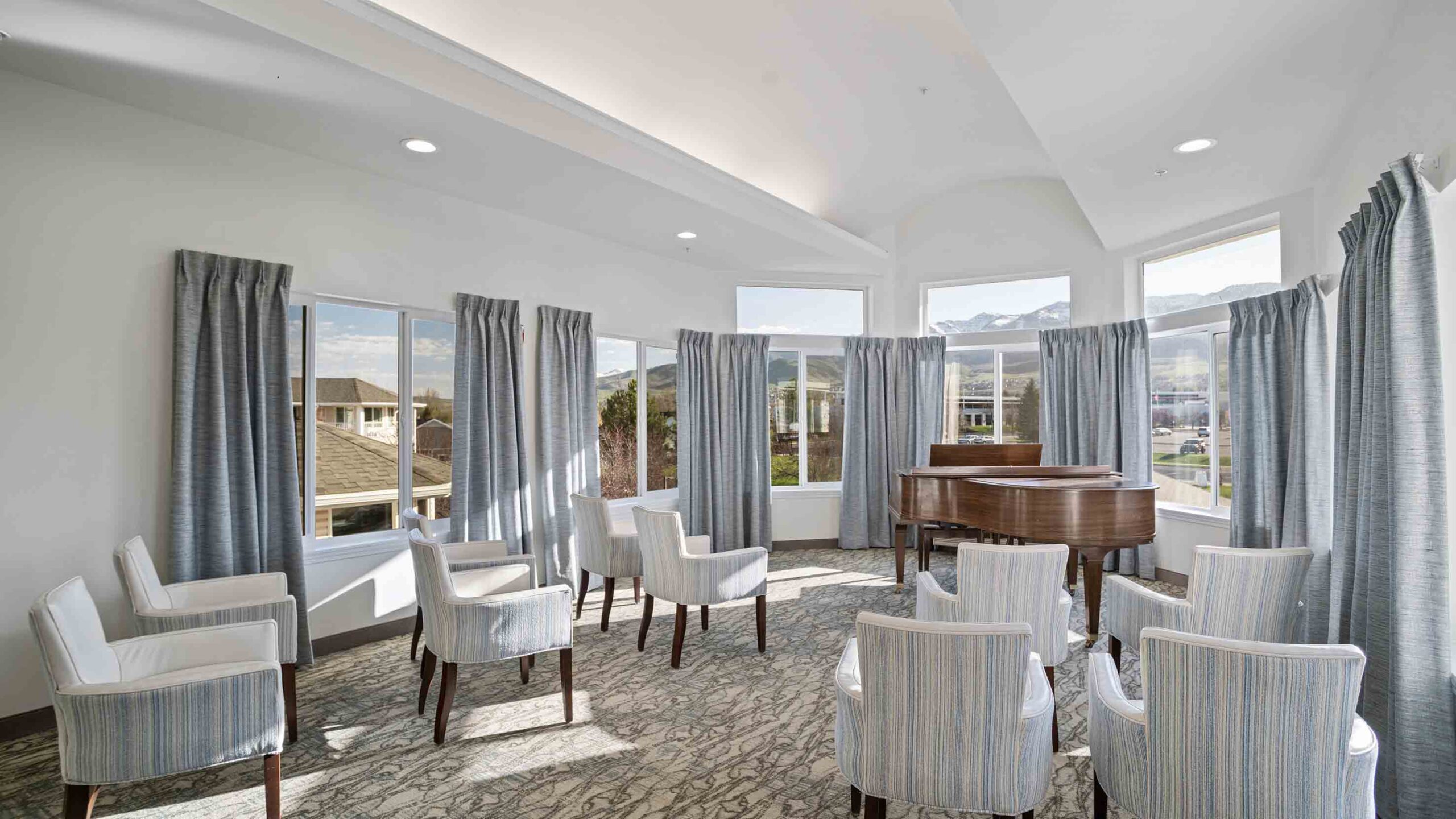 A bright, cozy room with large windows and light gray curtains. There are nine white and gray striped armchairs arranged in a semi-circle, and a grand piano in the corner with a bench. The carpet has a subtle gray pattern. The room is well-lit and inviting.