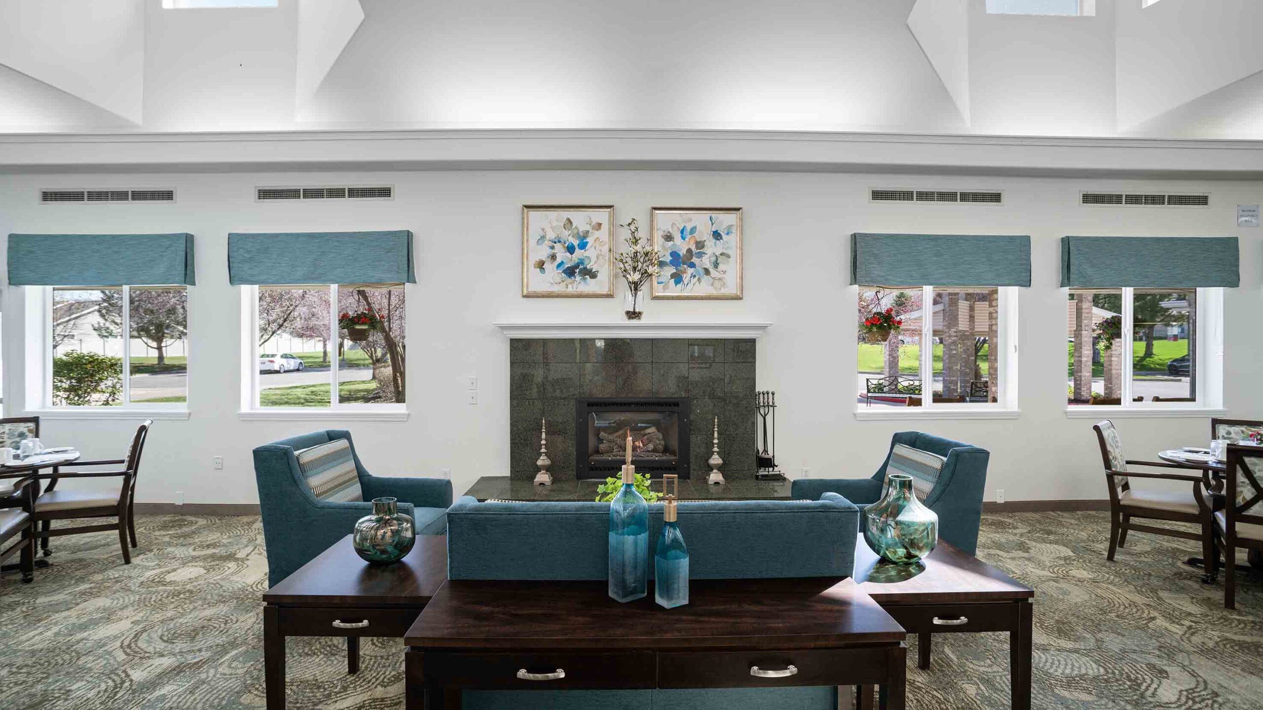 A well-lit living room with a fireplace, flanked by two blue armchairs and symmetrical windows. Two botanical paintings hang above the fireplace. A wooden coffee table with decorative blue vases stands in the foreground.