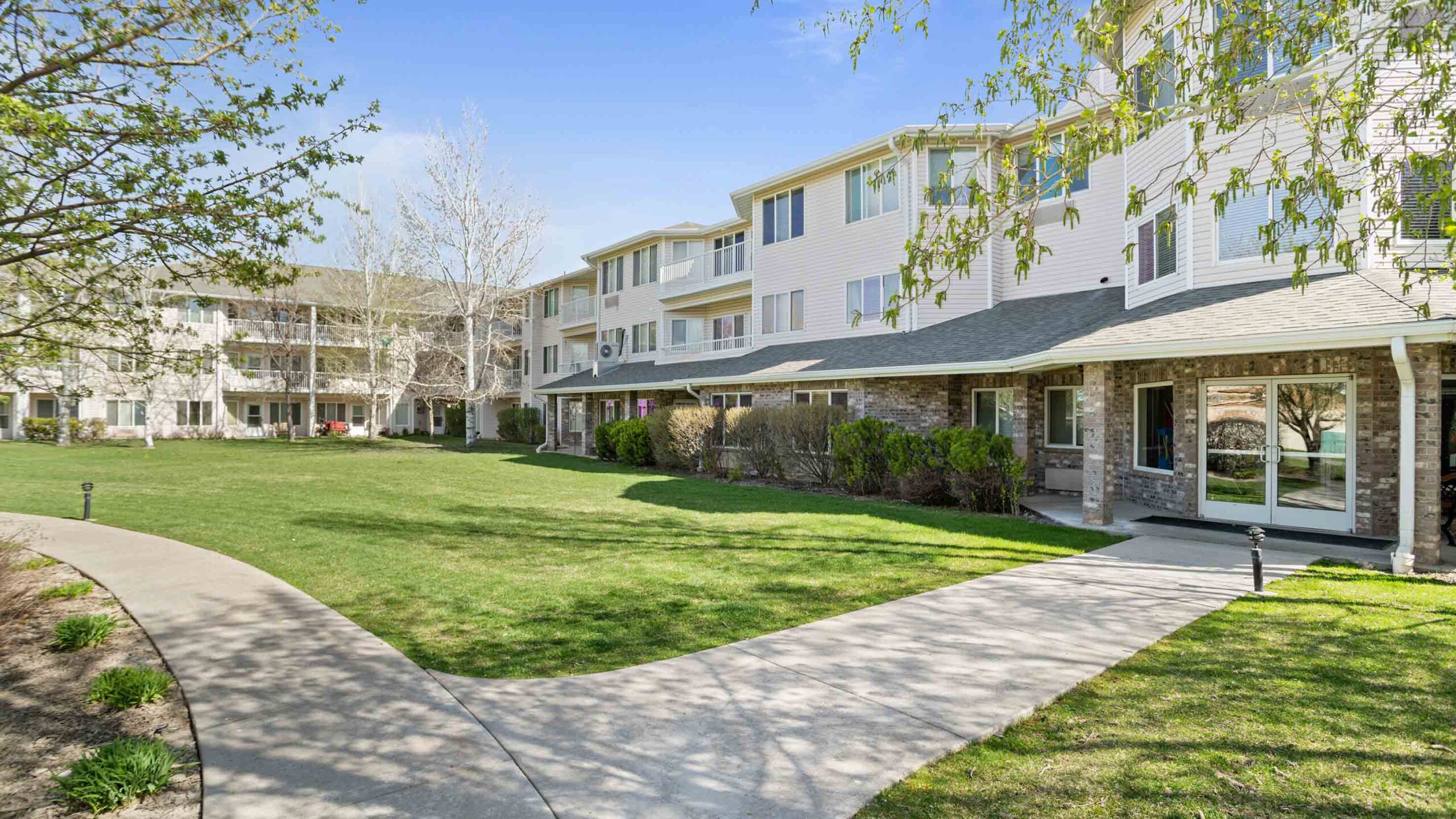 A well-maintained, multi-story residential building with white siding, surrounded by neatly trimmed bushes and a lush green lawn. A concrete walkway curves through the lawn, leading to the entrances. Trees with early spring leaves frame the scene under a clear blue sky.