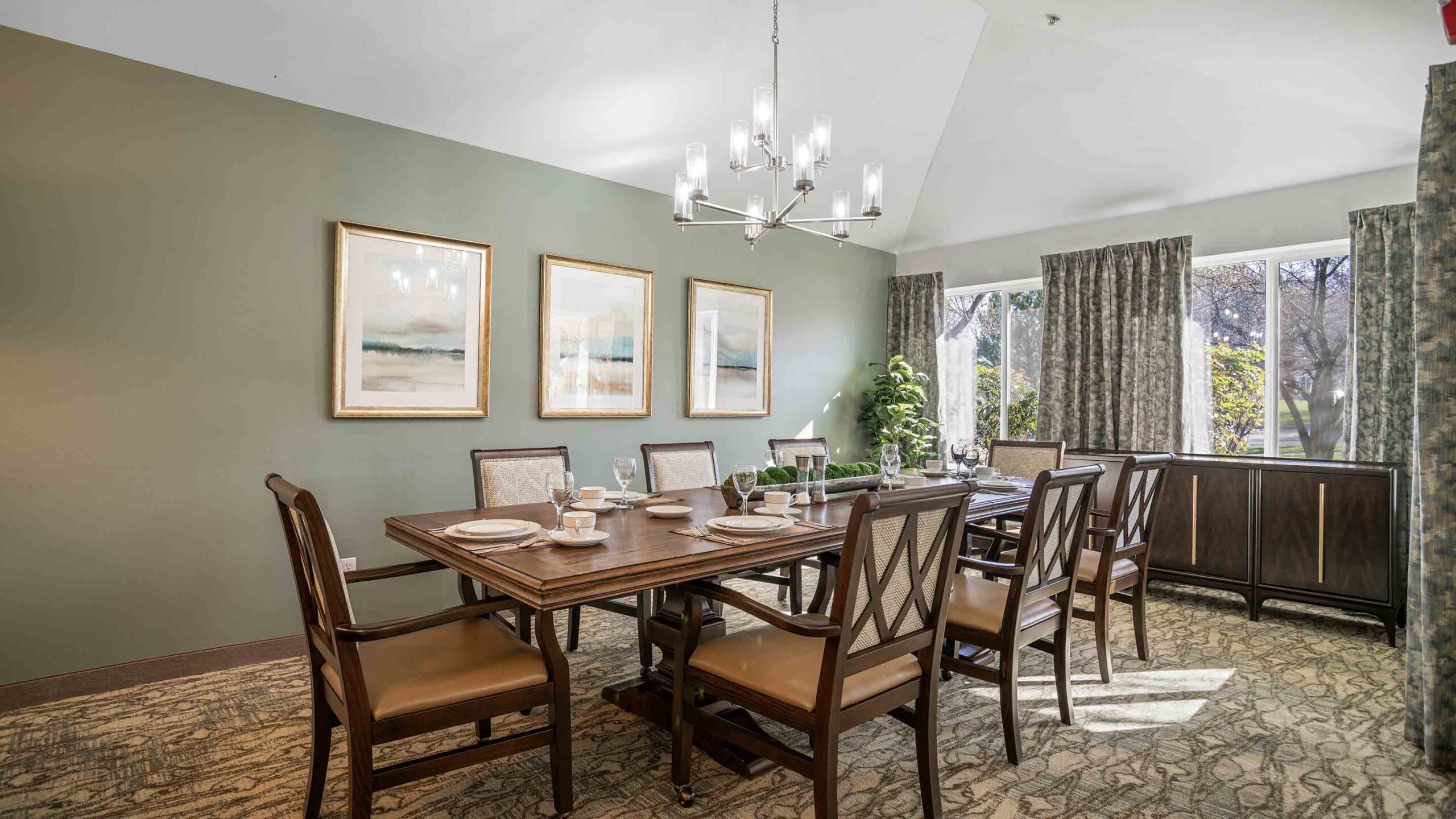 A dining room with a large wooden table and eight chairs. The table is set with white plates, glasses, and napkins. Three landscape paintings hang on the green wall. A modern chandelier is above the table. Large windows with curtains allow natural light in.