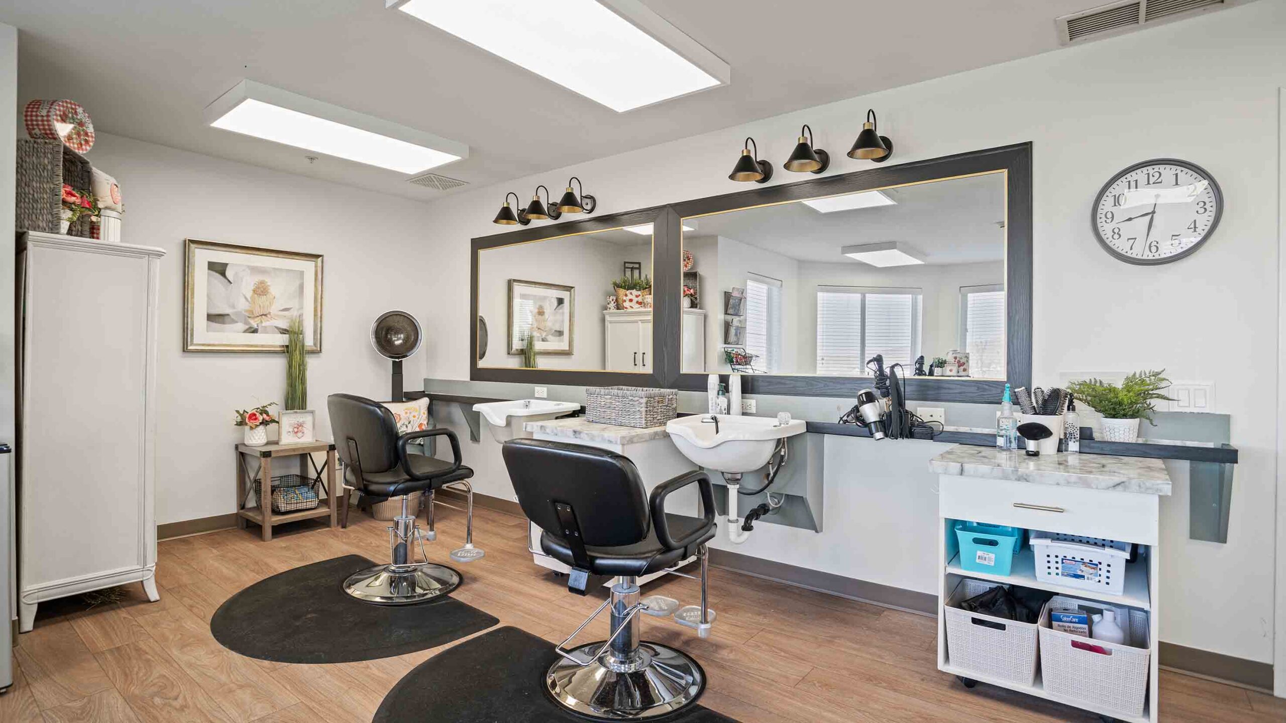 A modern hair salon features two styling stations with black chairs, large mirrors, and overhead lights. The room is decorated with framed art, a wall clock, and various beauty supplies on shelves and counters. The floor is wooden, creating a warm atmosphere.