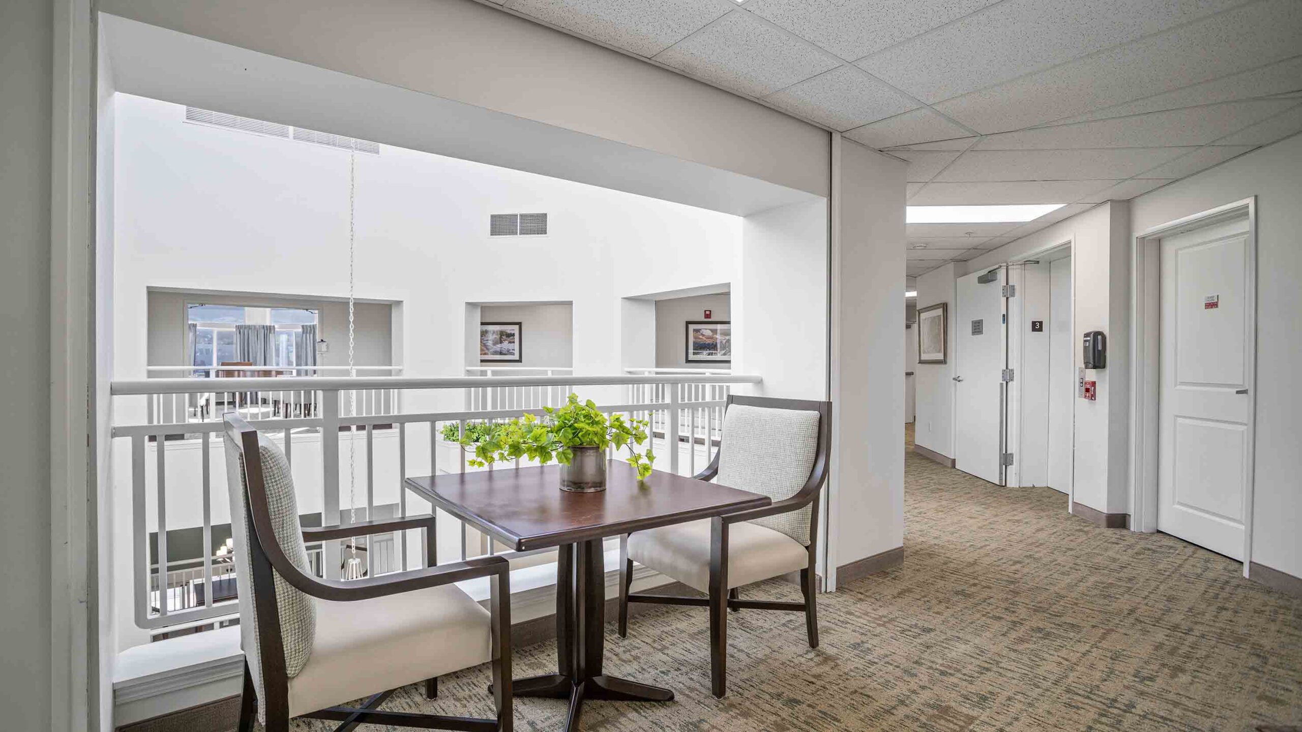 A quiet, brightly lit indoor atrium with a small table and two cushioned chairs next to a white railing. The area features a potted plant on the table, neutral-toned carpet, white walls, and doors leading to rooms in the background.