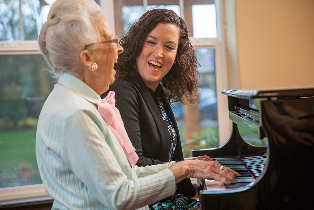 A senior woman and a younger woman are sitting at a piano, smiling and laughing together. The senior woman is playing the piano, while the younger woman watches enthusiastically. They are in a bright, cozy room with large windows in the background.