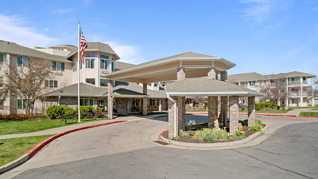 A modern, multi-story residential building with a covered entrance and a circular driveway, landscaped with grass and flowers. An American flag is raised on a flagpole near the entrance. The sky is clear and blue.