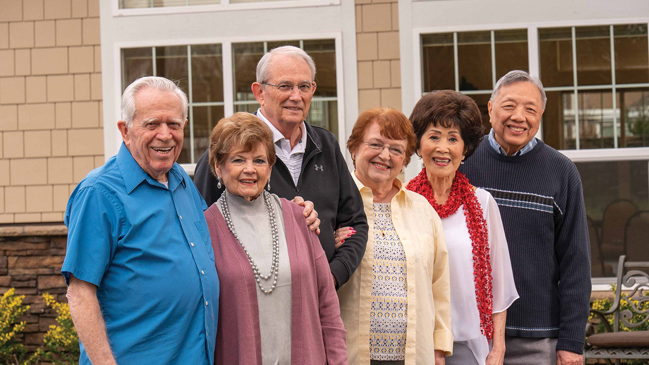 Six smiling older adults stand closely together outside in front of a building with large windows, enjoying each other's company on a pleasant day.
