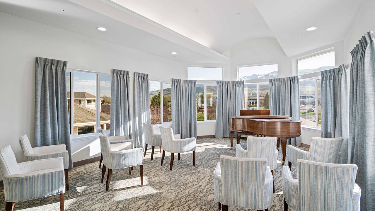 A bright room with large windows, blue-gray curtains, eight upholstered chairs arranged in a semi-circle, and a grand piano in the corner. Natural light fills the space, highlighting light carpet and white walls.