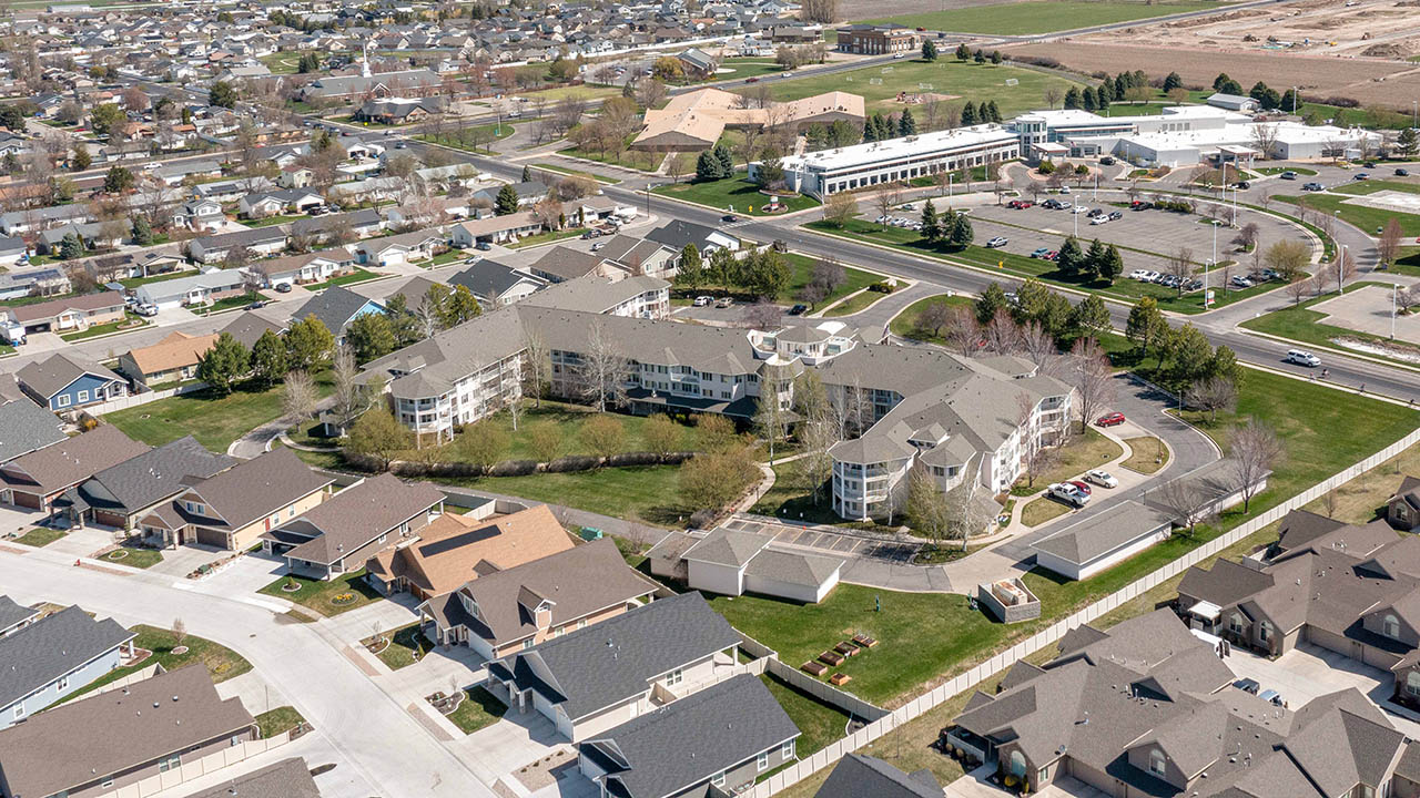 Aerial view of a residential neighborhood with houses, roads, and a large multi-story apartment complex surrounded by trees, lawns, and parking areas. Nearby, schools and open fields are visible.