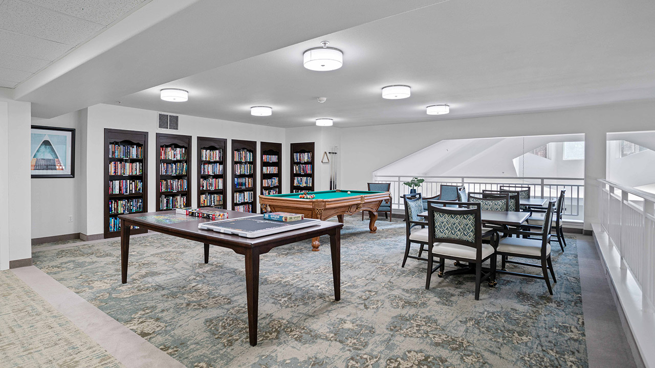 A bright game room features a pool table, a table with board games, a table with chairs, bookshelves filled with books, light fixtures on the ceiling, and a large window letting in natural light.
