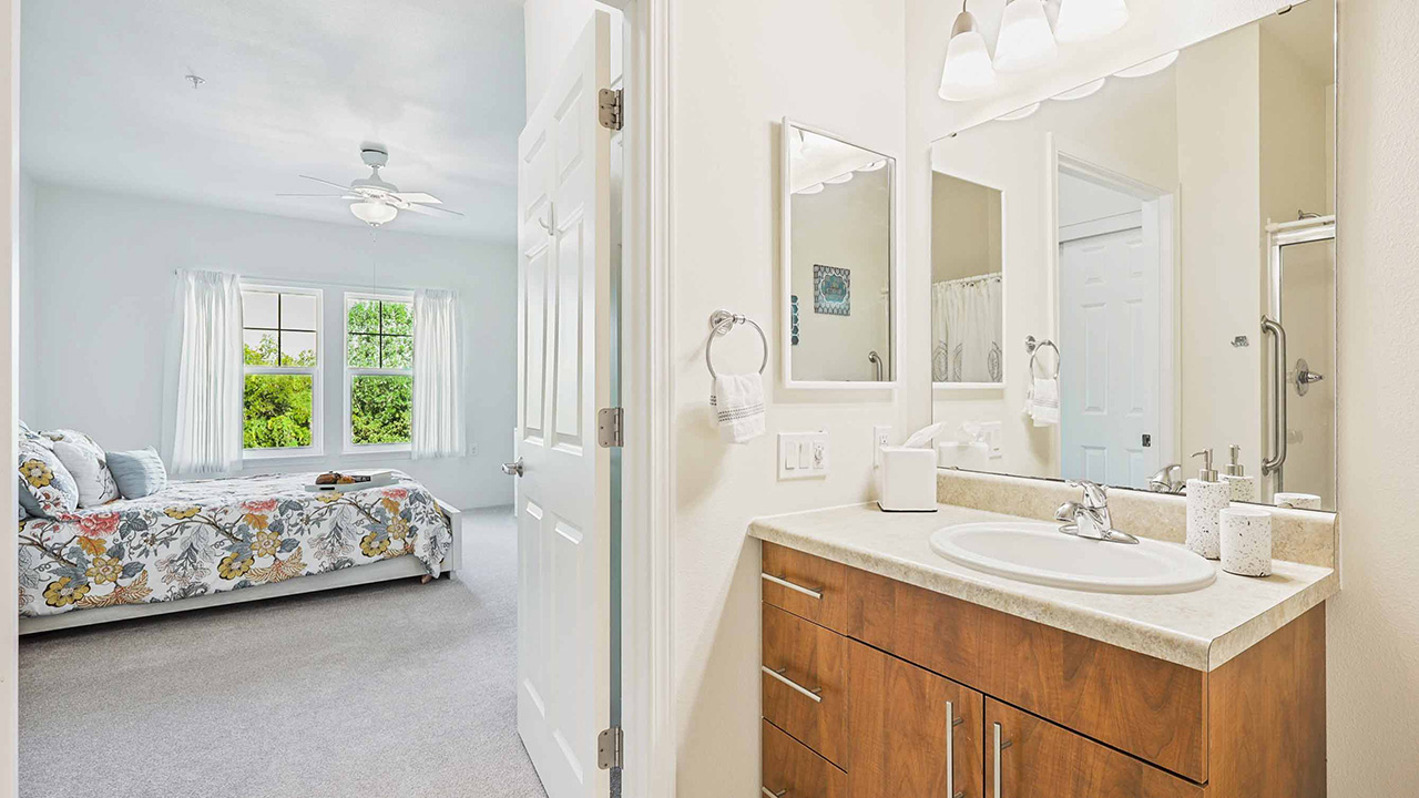 A bright bathroom with a wooden vanity and a white sink is adjacent to a bedroom with a floral bedspread, large windows, white curtains, and neutral walls and carpet. Natural light fills both rooms.