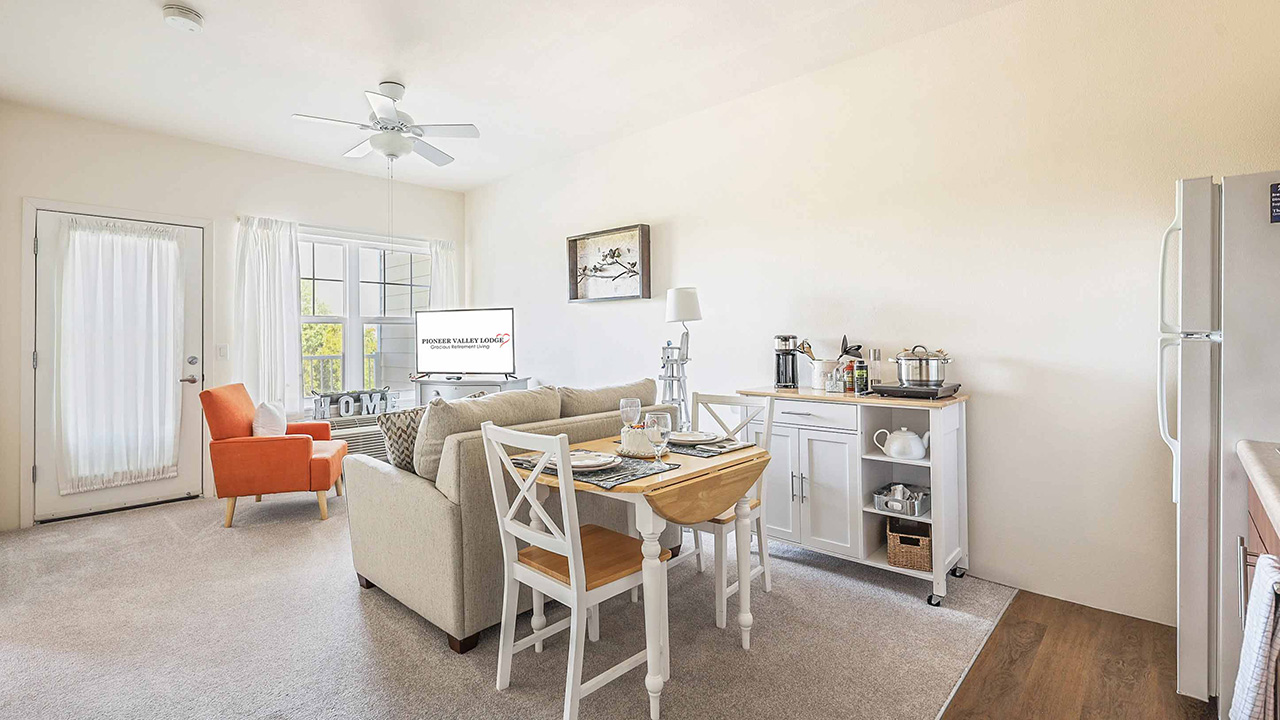 Bright, cozy living and dining area with a beige sofa, orange armchair, dining table set for two, small kitchen cart, and a TV by the window; white walls and large windows let in natural light.