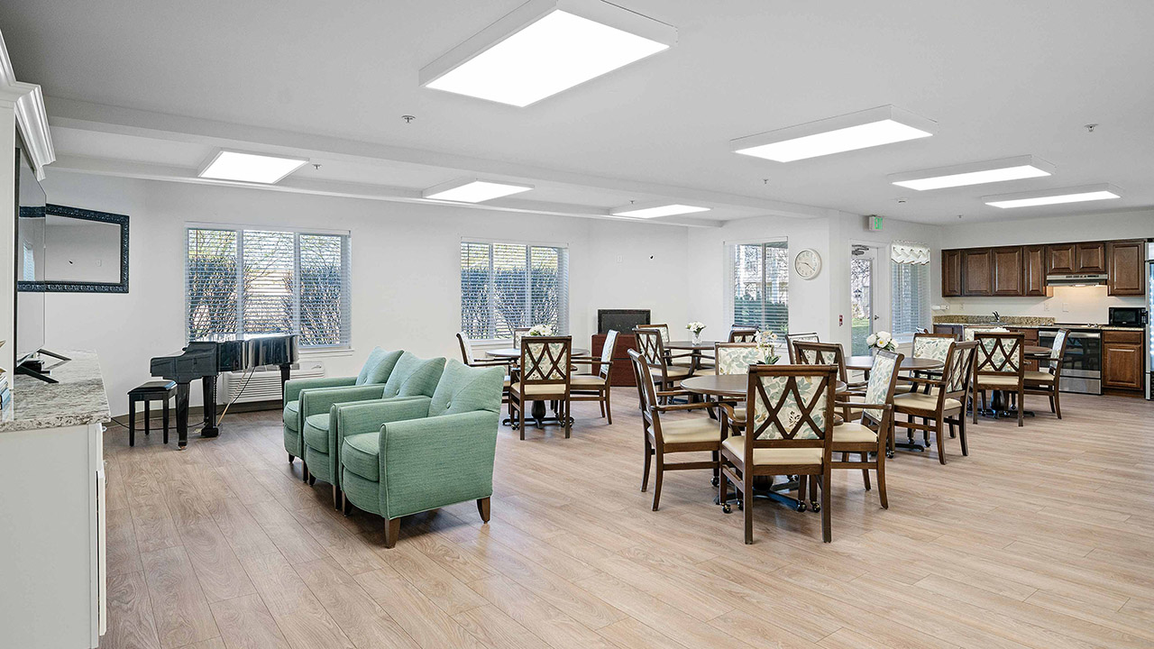 Spacious, bright room with wooden floors, several dining tables and chairs, green armchairs, a black piano, and a kitchen area in the background; large windows let in natural light.