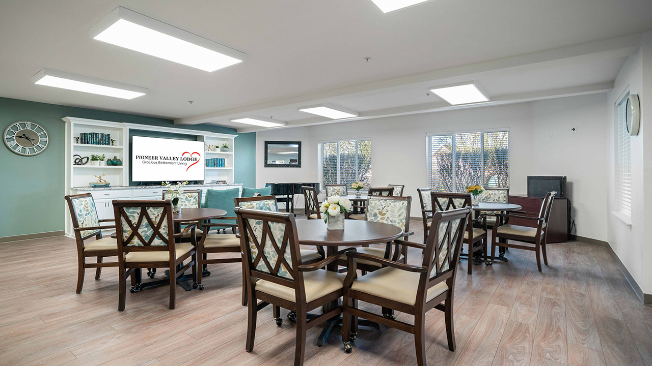 A bright, spacious room with several round tables and chairs, light wood floors, large windows, and a wall-mounted TV. Decorative shelves and a clock adorn the back wall. The space appears to be a communal area.