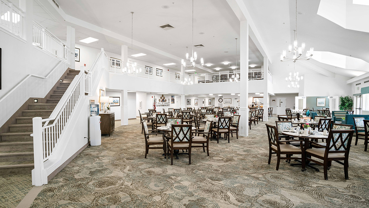 Spacious, well-lit dining room with neatly arranged tables and chairs, neutral decor, chandeliers, a staircase on the left, and a balcony area above.