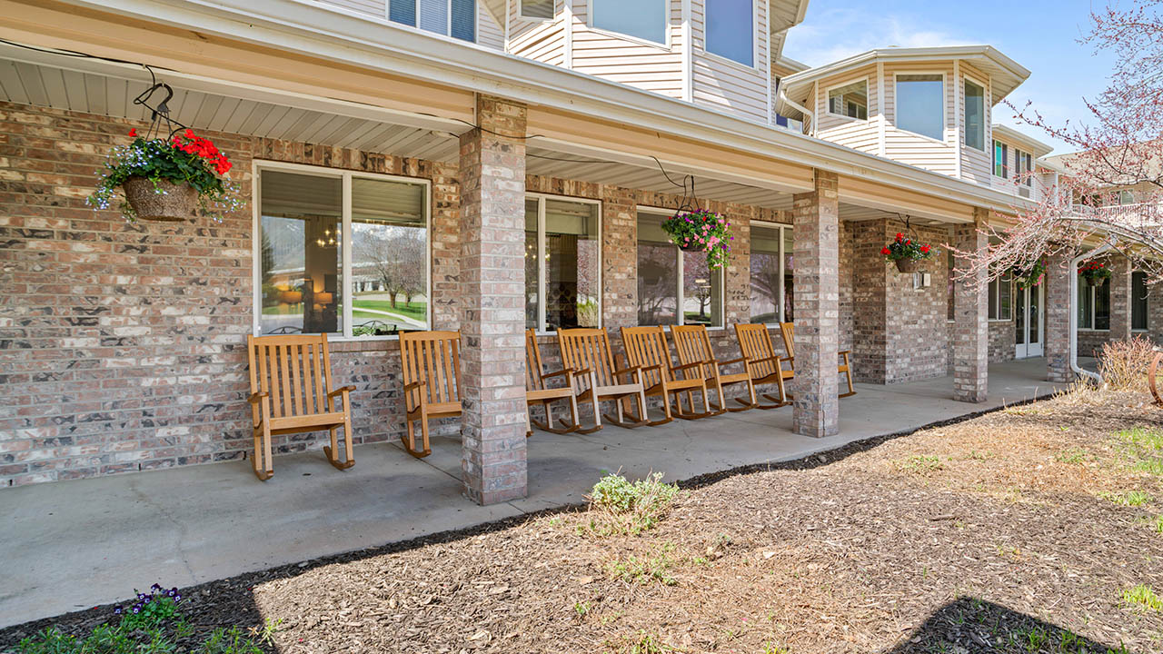 A row of wooden rocking chairs sits on a covered porch outside a brick building, with hanging flower baskets and large windows reflecting trees and greenery.
