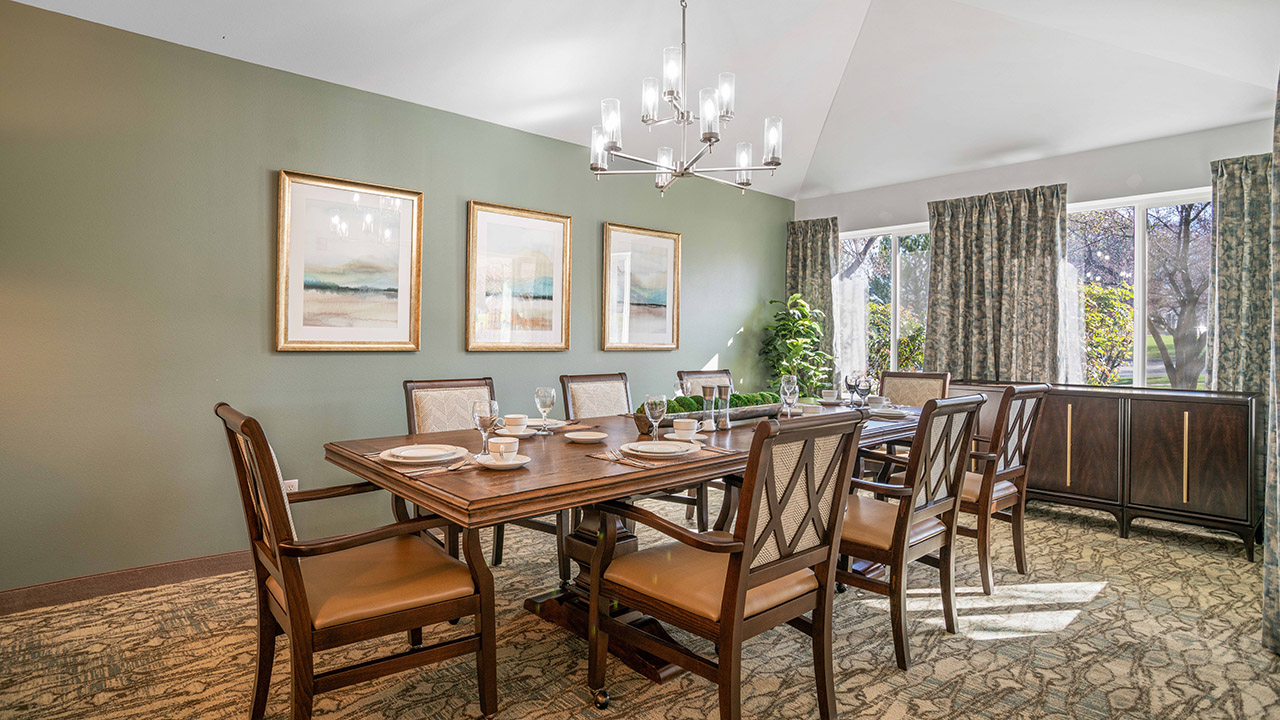 Elegant dining room with a wooden table set for six, matching chairs, three framed artworks on a green wall, large windows with patterned curtains, and a modern chandelier overhead. Sunlight brightens the space.