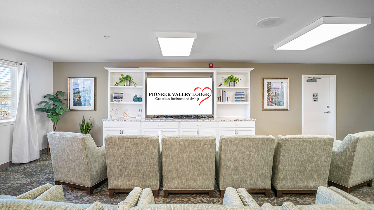 A bright, modern lounge with rows of cushioned chairs facing a TV displaying "Pioneer Valley Lodge: Gracious Retirement Living." Shelves with books, plants, and decor are on either side of the TV.