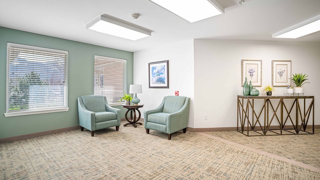 A bright, modern sitting area with two light blue armchairs, a round side table with a lamp, and framed art on the wall. Nearby, a console table holds plants, vases, and framed pictures. Large windows let in natural light.