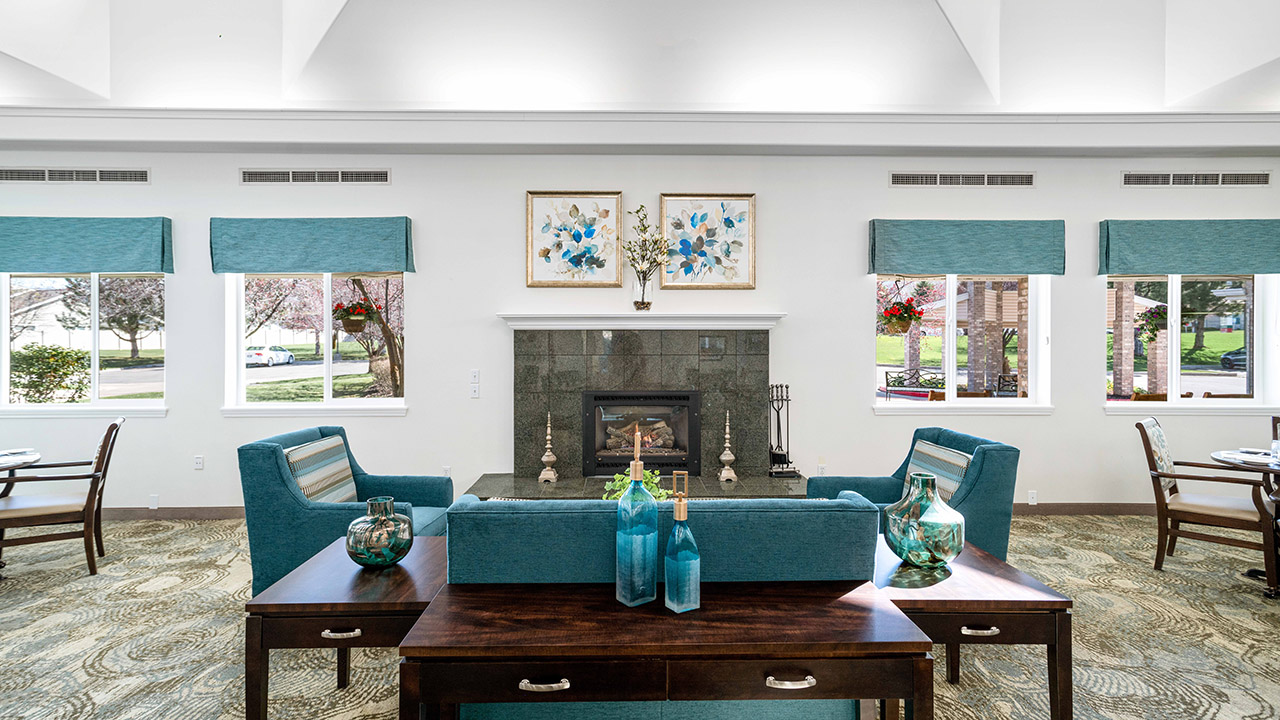 A bright living room with teal armchairs and a sofa facing a fireplace, decorative vases on a wooden table, two floral artworks above the mantel, and large windows letting in natural light.