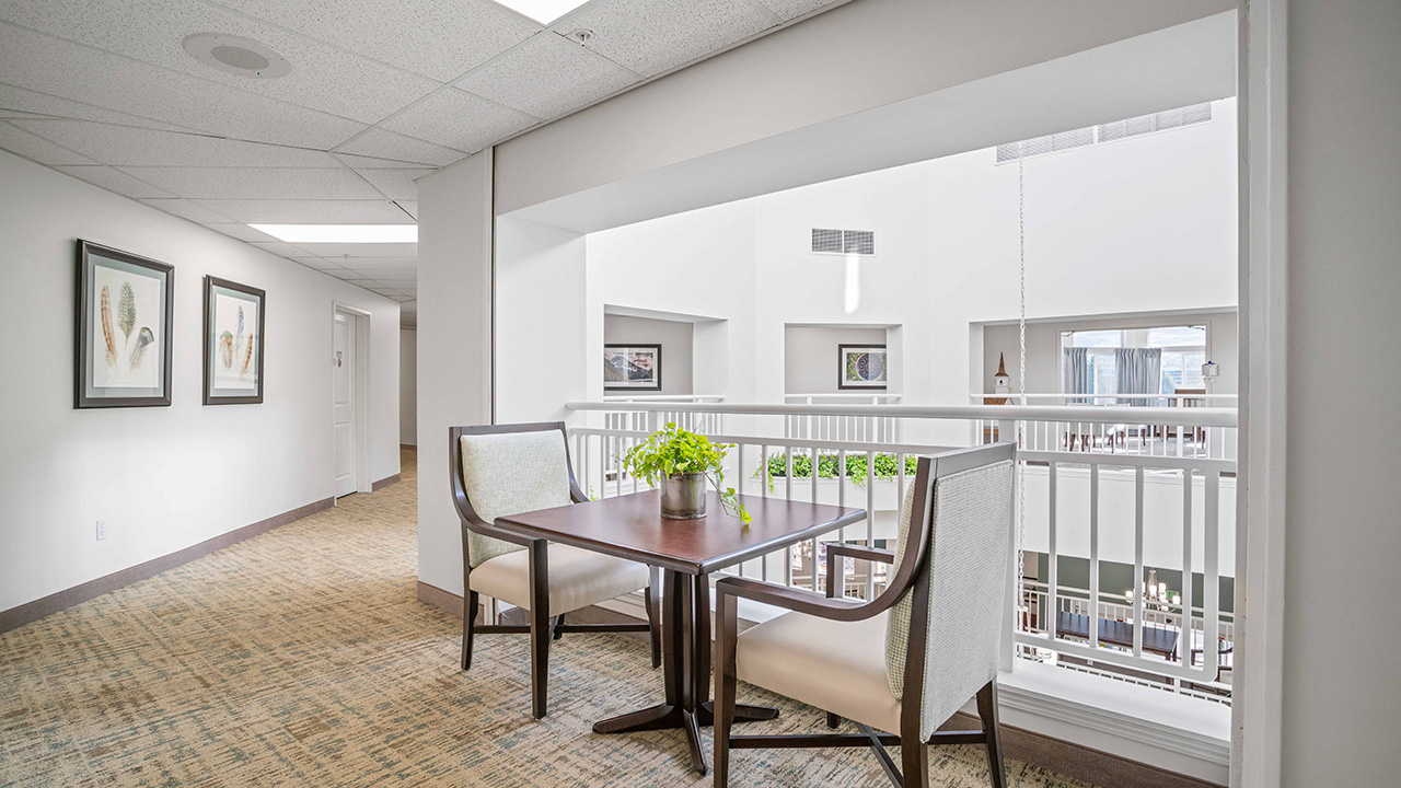 A small table with two chairs and a potted plant sits near a white railing in a bright hallway with beige carpet and framed artwork on the walls. Large windows let in natural light from above.