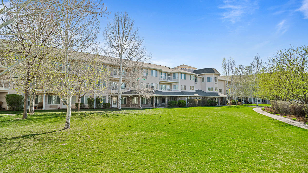 A large, light-colored apartment building with multiple balconies overlooks a spacious, well-maintained green lawn bordered by trees and a curved walking path under a clear blue sky.