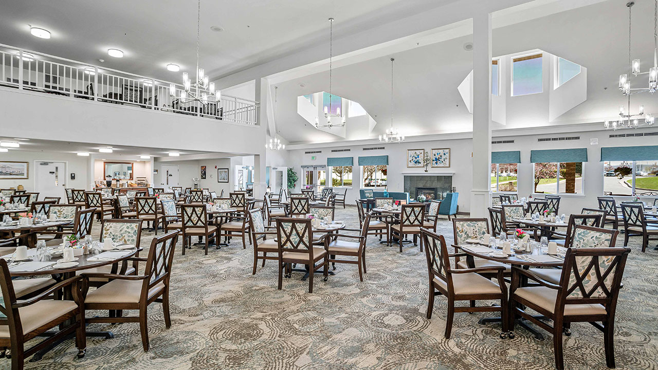 A spacious, modern dining room with high ceilings, chandeliers, large windows, and neatly arranged wooden tables and chairs set for a meal. The room has light-colored decor and a carpeted floor with a subtle pattern.