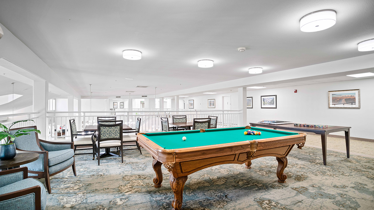 A bright, modern game room with a green felt pool table, billiard balls, cue sticks, a card table, chairs, board games, and artwork on the white walls, with large windows allowing natural light in.