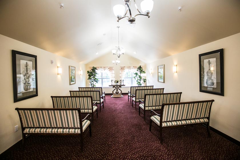 A small chapel with a vaulted ceiling and maroon carpet is furnished with striped benches arranged in rows. There are framed pictures and wall lights on the cream-colored walls, plants near the windows at the far end, and a small table in the center.