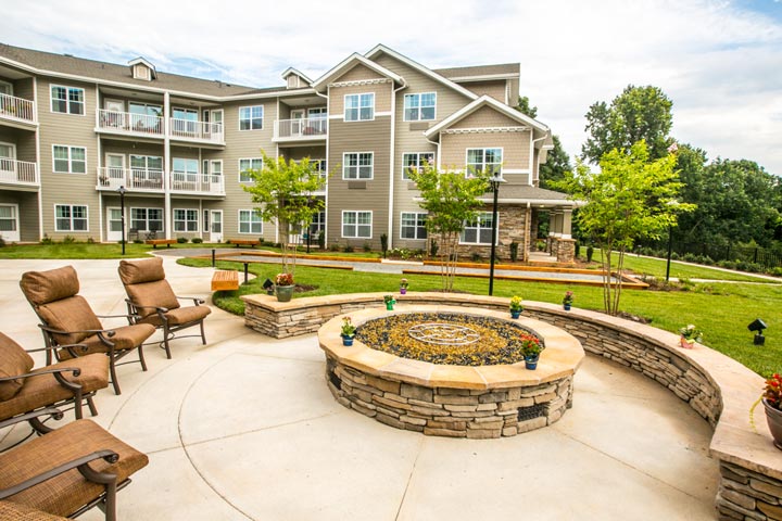 A landscaped outdoor area with a circular stone fire pit surrounded by chairs. Behind the seating area, there are well-maintained lawns, young trees, and a large, multi-story apartment building with balconies and large windows. The sky is partly cloudy.