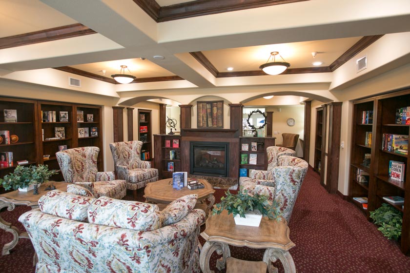 A cozy room featuring patterned armchairs arranged around wooden tables. Shelves filled with books line the walls, and a fireplace with a mantle is at the center. The room is well-lit with elegant ceiling lights and has a rich, red-carpeted floor.