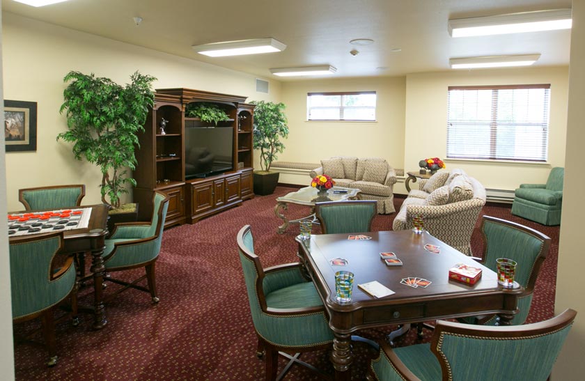 A cozy community room with maroon carpet, green cushioned chairs, and beige sofas. There are tables with checkerboards and card games, a large TV in a wooden entertainment center, and potted plants decorating the space. Two windows provide natural light.