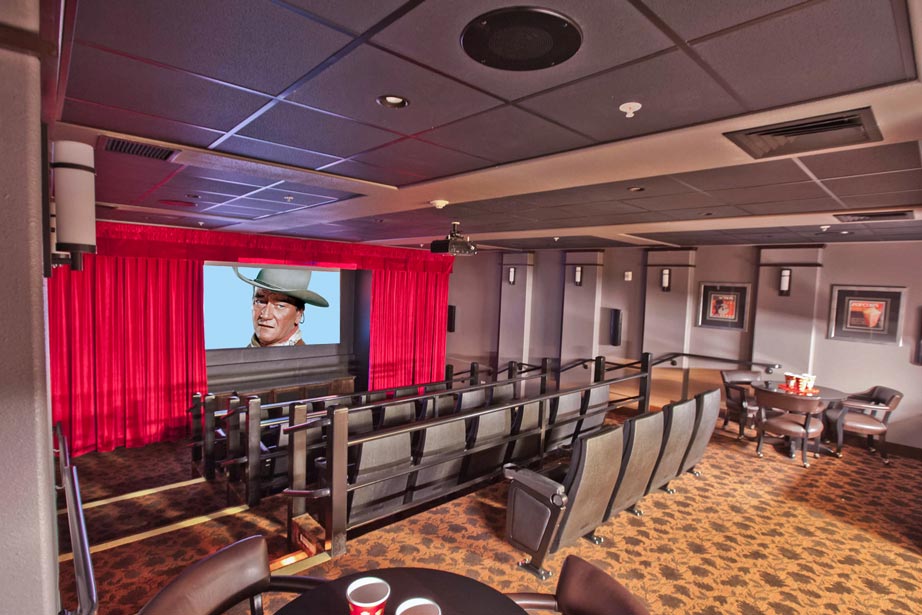 A home theater room featuring tiered seating with leather recliners, a large screen displaying a classic film scene, red curtains framing the screen, modern lighting on the ceiling, and a small table with refreshments in the foreground. The decor is cozy and inviting.