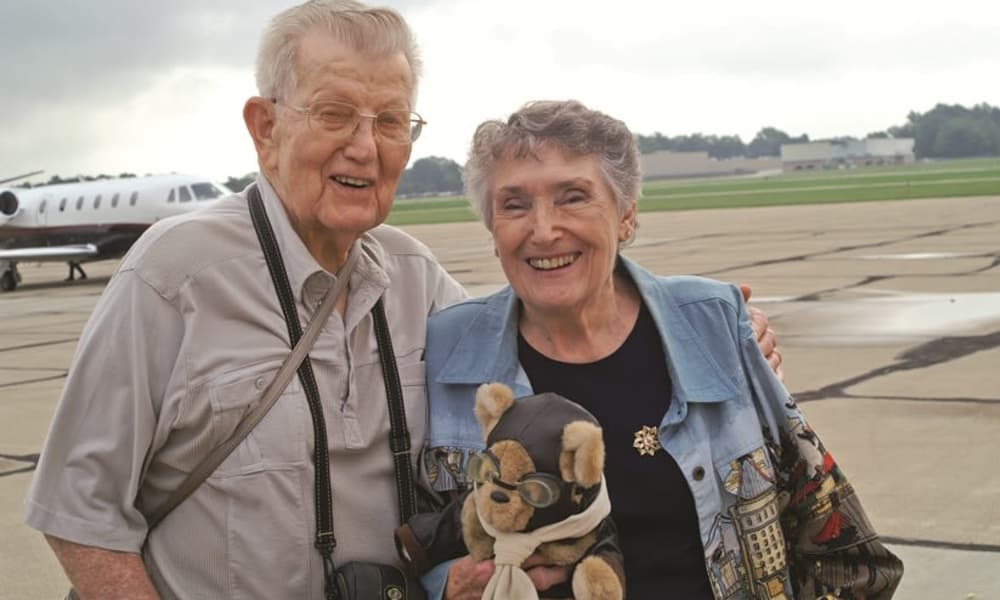 A smiling elderly couple stands on an airport tarmac. The man is on the left wearing glasses, a light-colored shirt, and a camera around his neck. The woman, to his right, holds a stuffed animal dressed as a pilot. An airplane is visible in the background.
