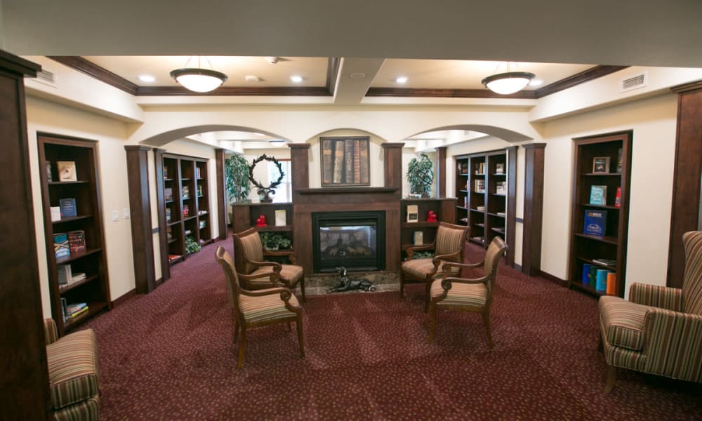 A cozy room with maroon carpet featuring a fireplace surrounded by bookshelves filled with books and decorative items. Four cushioned, striped armchairs are arranged around a small central table. The room is softly lit by ceiling lights.