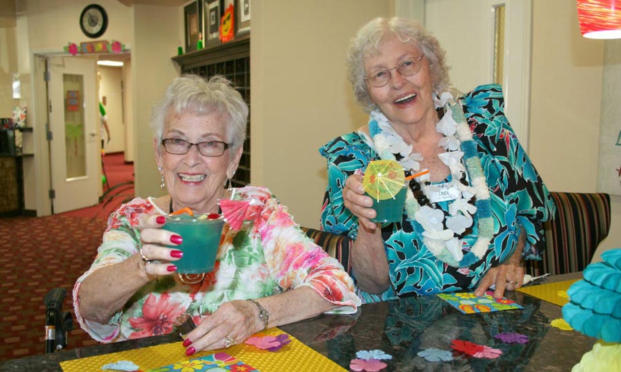 Two elderly women sit at a table enjoying tropical drinks with colorful garnishes. Both women are smiling and wearing floral prints; one wears a lei. The setting appears festive with vibrant decorations and a laid-back ambiance.