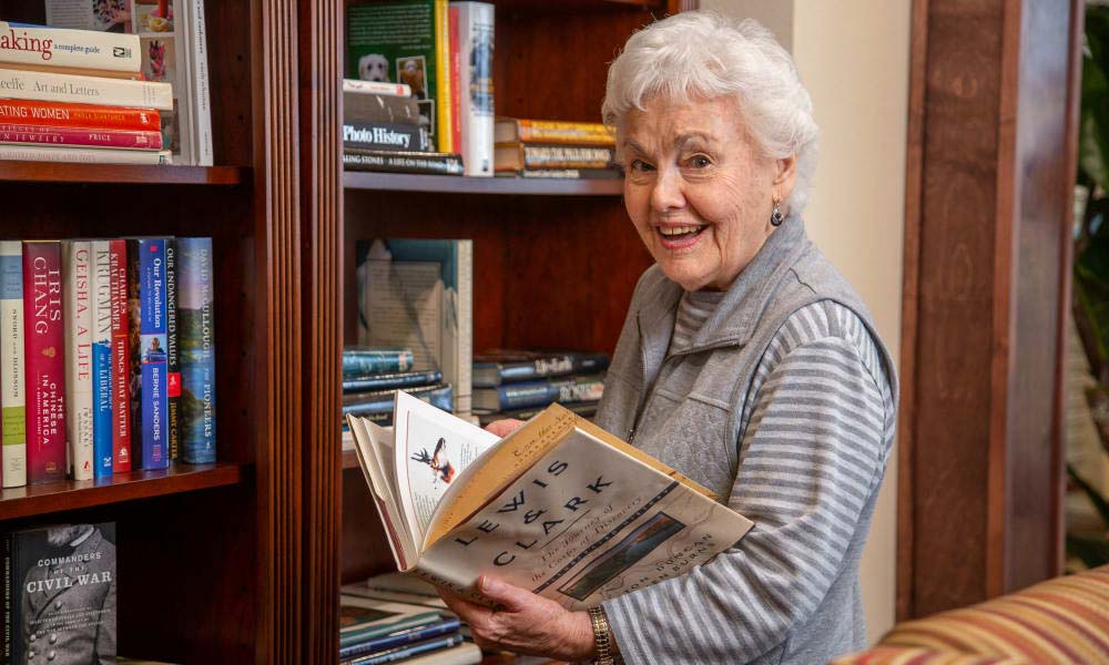 An elderly woman with short white hair and a gray jacket stands in front of a bookshelf filled with various books. She smiles as she holds an open book titled 