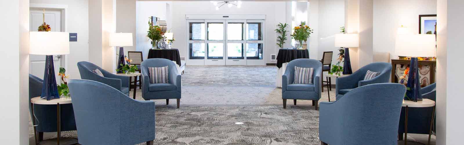 A spacious lobby with blue armchairs and tables, decorated with lamps and green plants. Large windows at the far end provide natural light, and patterned carpet covers the floor.