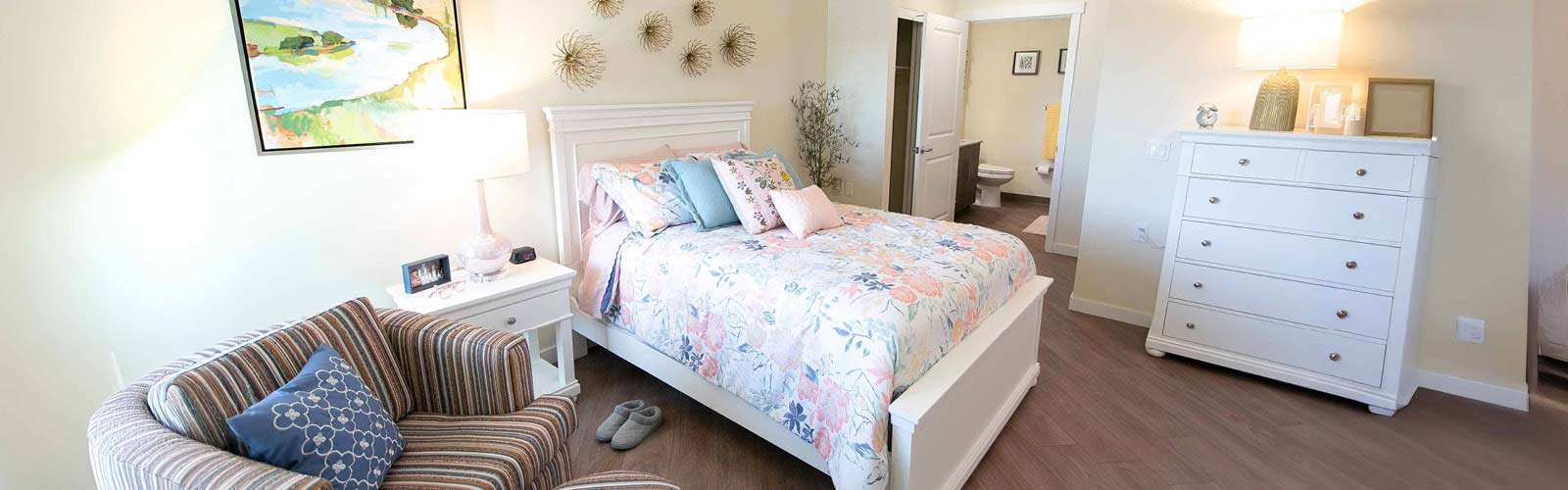 A cozy bedroom with a striped armchair, floral bedspread on a white bed, and a white dresser. Wall art and decorative pieces complement the soft lighting from a table lamp, creating a serene atmosphere. An open door leads to a bathroom.