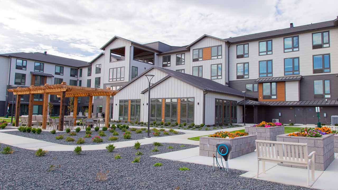 A modern multi-story building with large windows and a mix of gray and white exterior. The landscaped area in front features a wooden pergola, pathways, benches, and colorful flowerbeds. The sky is cloudy.