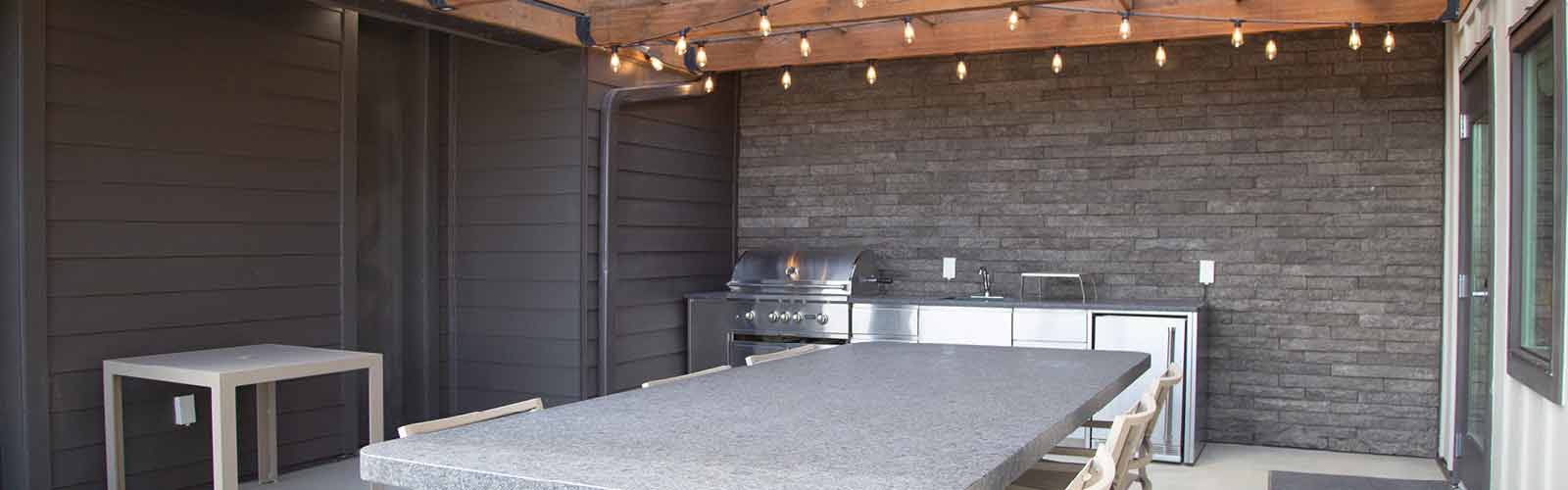 Outdoor patio with a long stone dining table, metal chairs, and a modern barbecue grill against a dark brick wall. String lights hang above, creating a cozy ambiance.