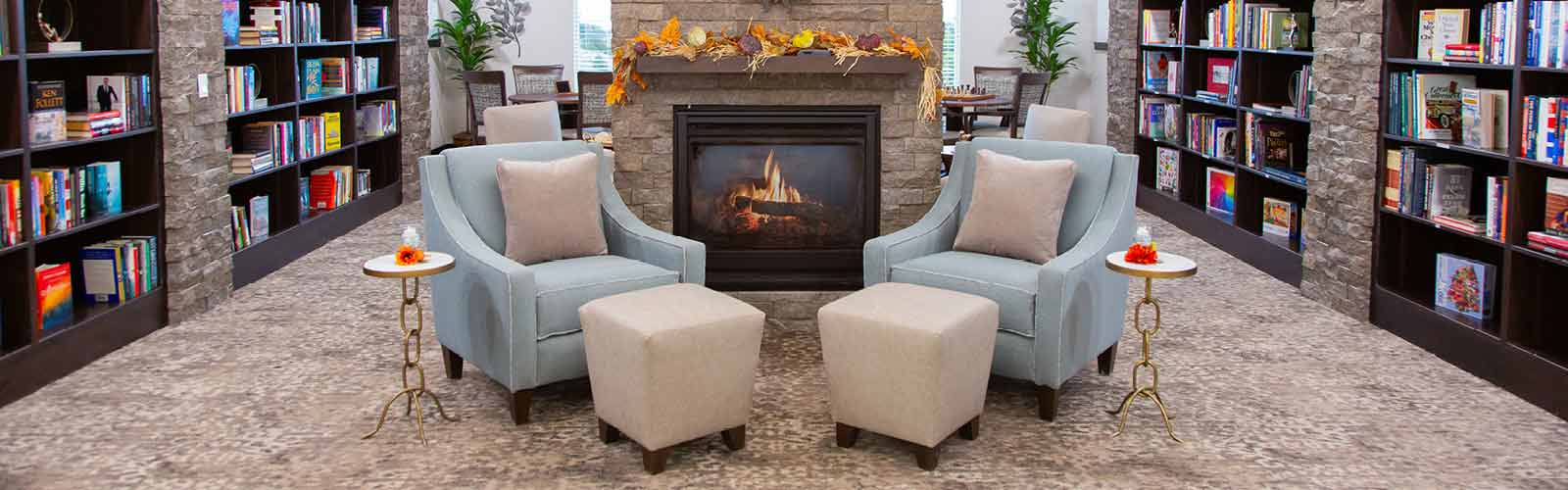 A cozy library with two blue armchairs and matching ottomans facing a stone fireplace. Shelves filled with books line the walls, and small tables with autumn decor sit beside the chairs. The warm ambiance is inviting and comfortable.