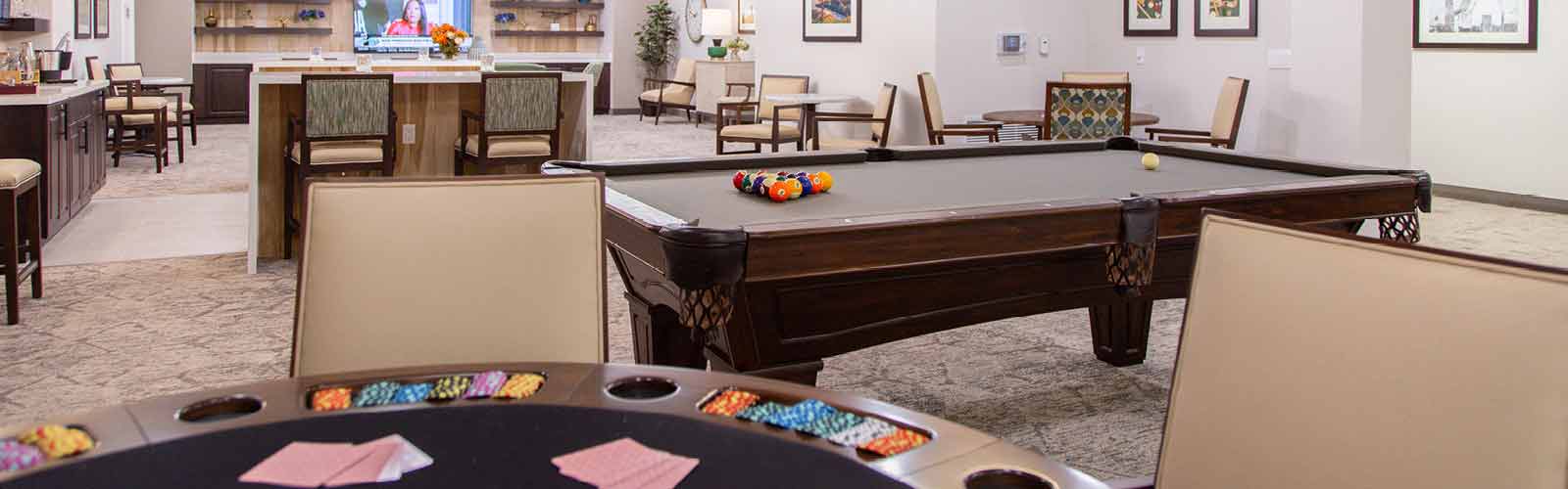 A recreational room with a pool table, poker table with chips and cards, and various seating arrangements. A TV is visible in the background, mounted on the wall, and the room is well-lit and carpeted.