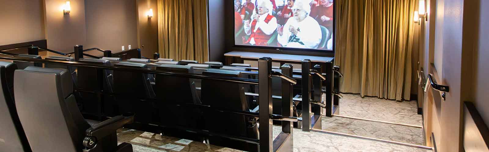 A small, dimly lit movie theater with tiered seating and comfortable black chairs. The screen displays a scene with people dressed in red and white costumes. The walls are adorned with sconces, and curtains frame the screen.