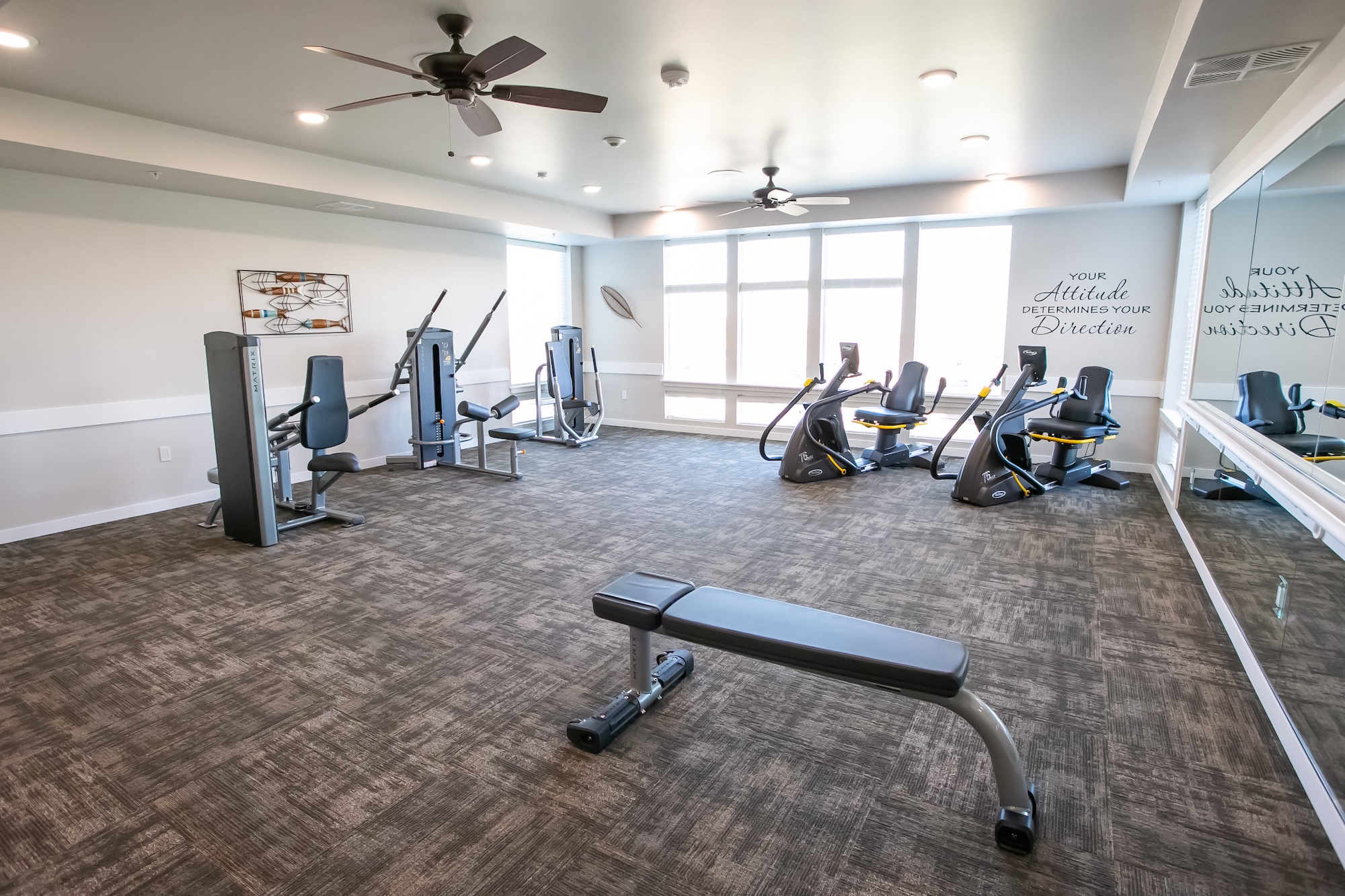 A modern gym with several exercise machines, including cardio and weight equipment, and motivational quotes on the wall. The room features large windows allowing natural light, ceiling fans, and a mirrored wall.